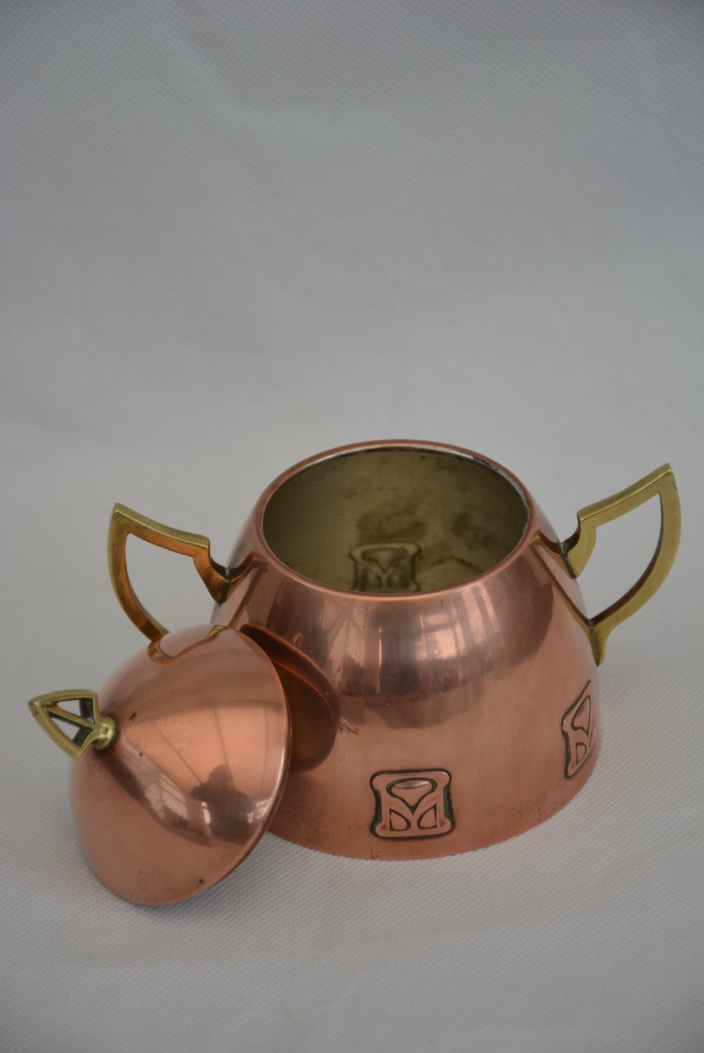 An Art Nouveau copper sugar bowl featuring angular brass handles, a conical lid with a stylised finial, and embossed geometric motifs around the base. View XI