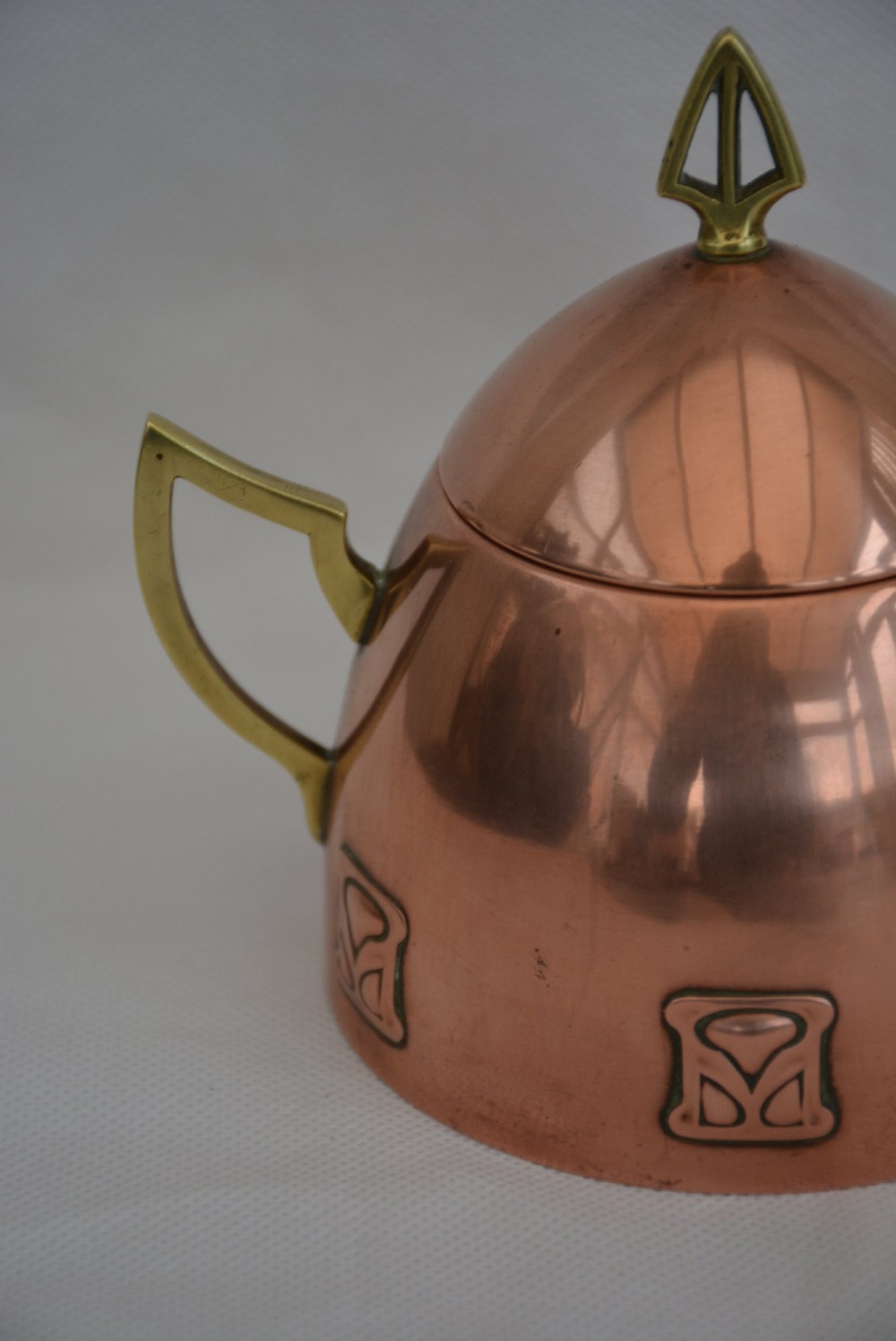 An Art Nouveau copper sugar bowl featuring angular brass handles, a conical lid with a stylised finial, and embossed geometric motifs around the base. View XIII