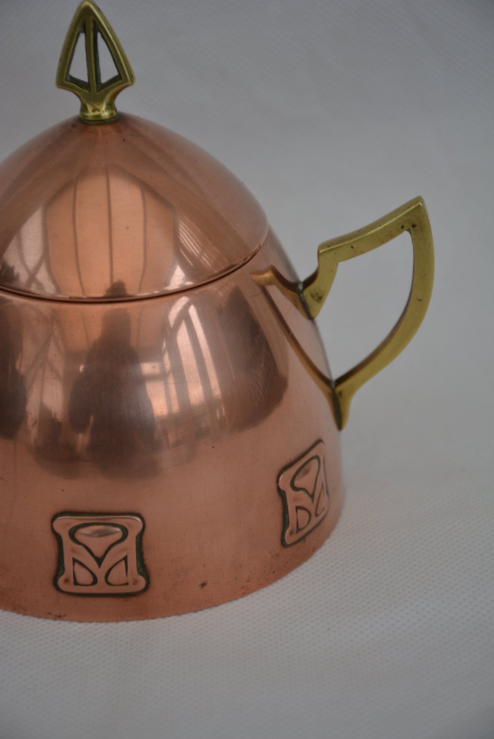 An Art Nouveau copper sugar bowl featuring angular brass handles, a conical lid with a stylised finial, and embossed geometric motifs around the base. View XIV