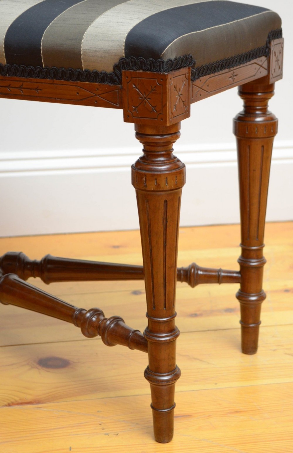 A Victorian walnut window seat featuring a striped upholstered top, fluted turned legs, and an intricate cross-stretcher with a central finial. View II