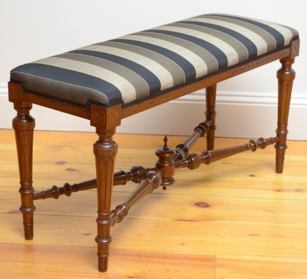A Victorian walnut window seat featuring a striped upholstered top, fluted turned legs, and an intricate cross-stretcher with a central finial. View V