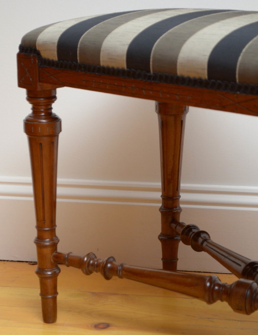 A Victorian walnut window seat featuring a striped upholstered top, fluted turned legs, and an intricate cross-stretcher with a central finial. View X