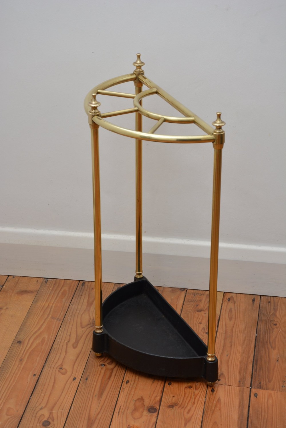 A semi-circular brass umbrella stand featuring finial tops, vertical tubular legs, a partitioned top frame, and a black drip tray base. View III