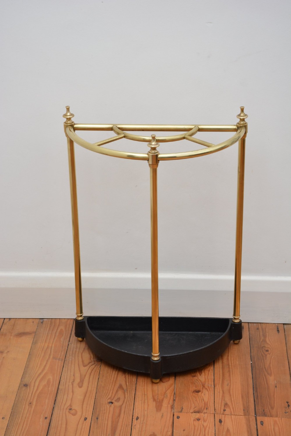 A semi-circular brass umbrella stand featuring finial tops, vertical tubular legs, a partitioned top frame, and a black drip tray base. View IV
