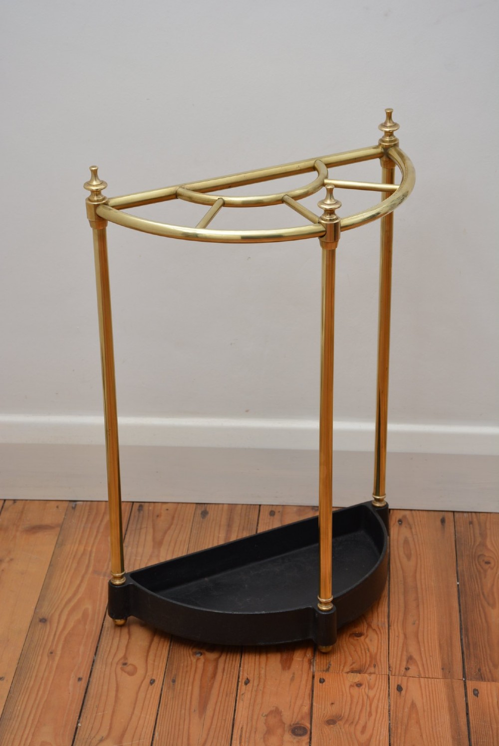 A semi-circular brass umbrella stand featuring finial tops, vertical tubular legs, a partitioned top frame, and a black drip tray base. View V