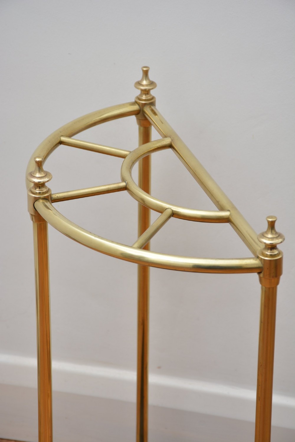A semi-circular brass umbrella stand featuring finial tops, vertical tubular legs, a partitioned top frame, and a black drip tray base. View VI