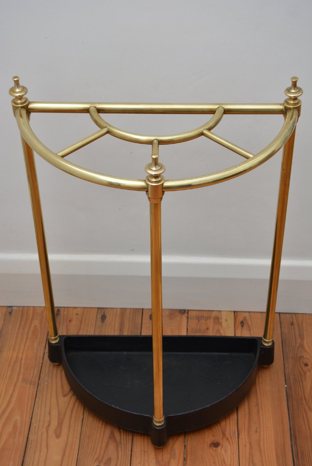 A semi-circular brass umbrella stand featuring finial tops, vertical tubular legs, a partitioned top frame, and a black drip tray base. View X