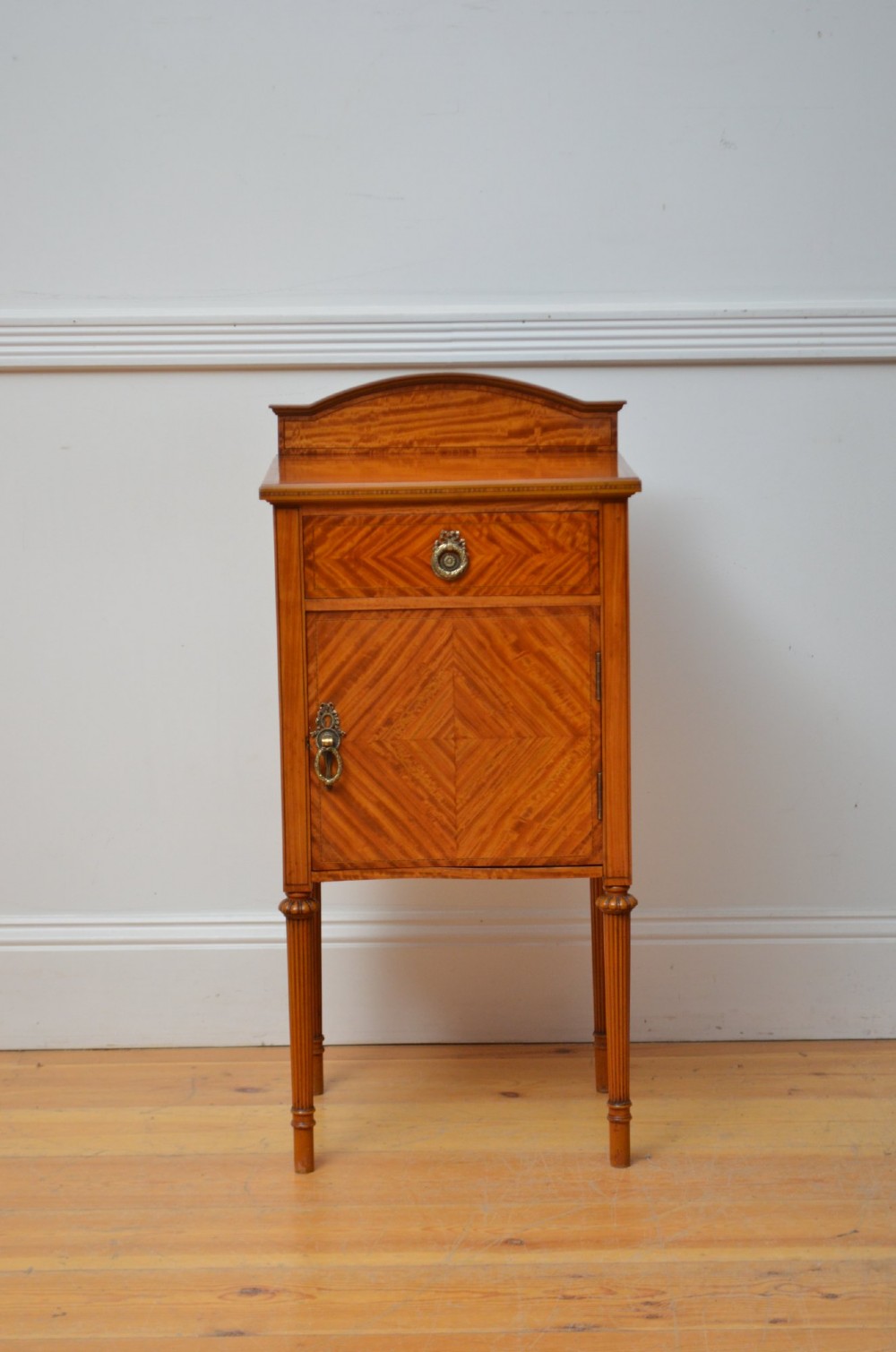 An Edwardian satinwood bedside cupboard featuring chevron veneer detailing, an upper drawer, a lower door, and turned reeded legs.