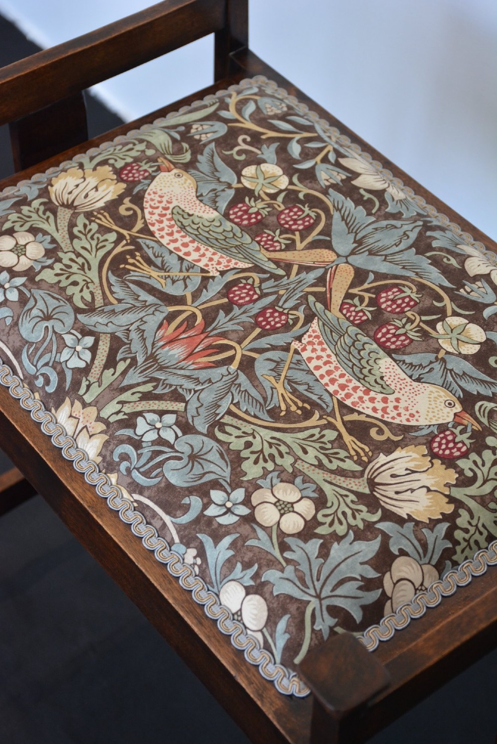 An antique dark mahogany Arts and Crafts stool featuring upright corner posts with square finials, vertical side splats, and a pattern-upholstered fabric seat. View II