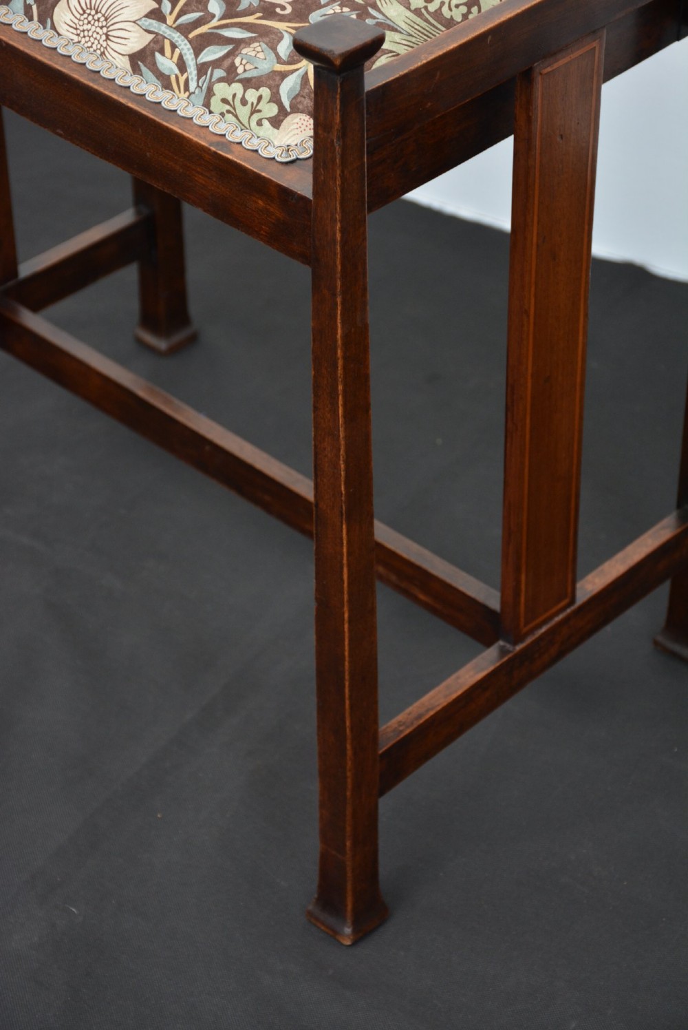 An antique dark mahogany Arts and Crafts stool featuring upright corner posts with square finials, vertical side splats, and a pattern-upholstered fabric seat. View VII