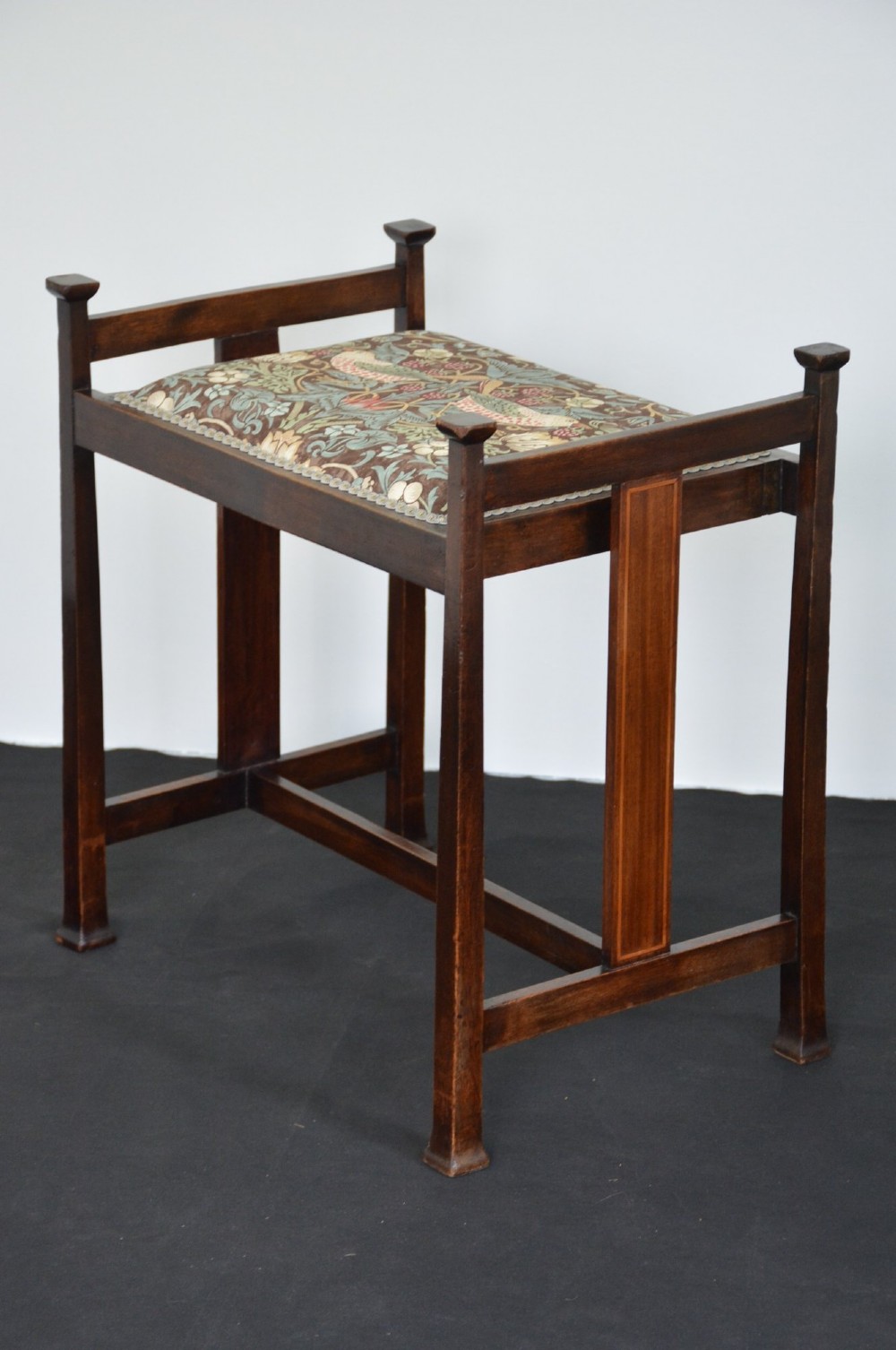 An antique dark mahogany Arts and Crafts stool featuring upright corner posts with square finials, vertical side splats, and a pattern-upholstered fabric seat. View X