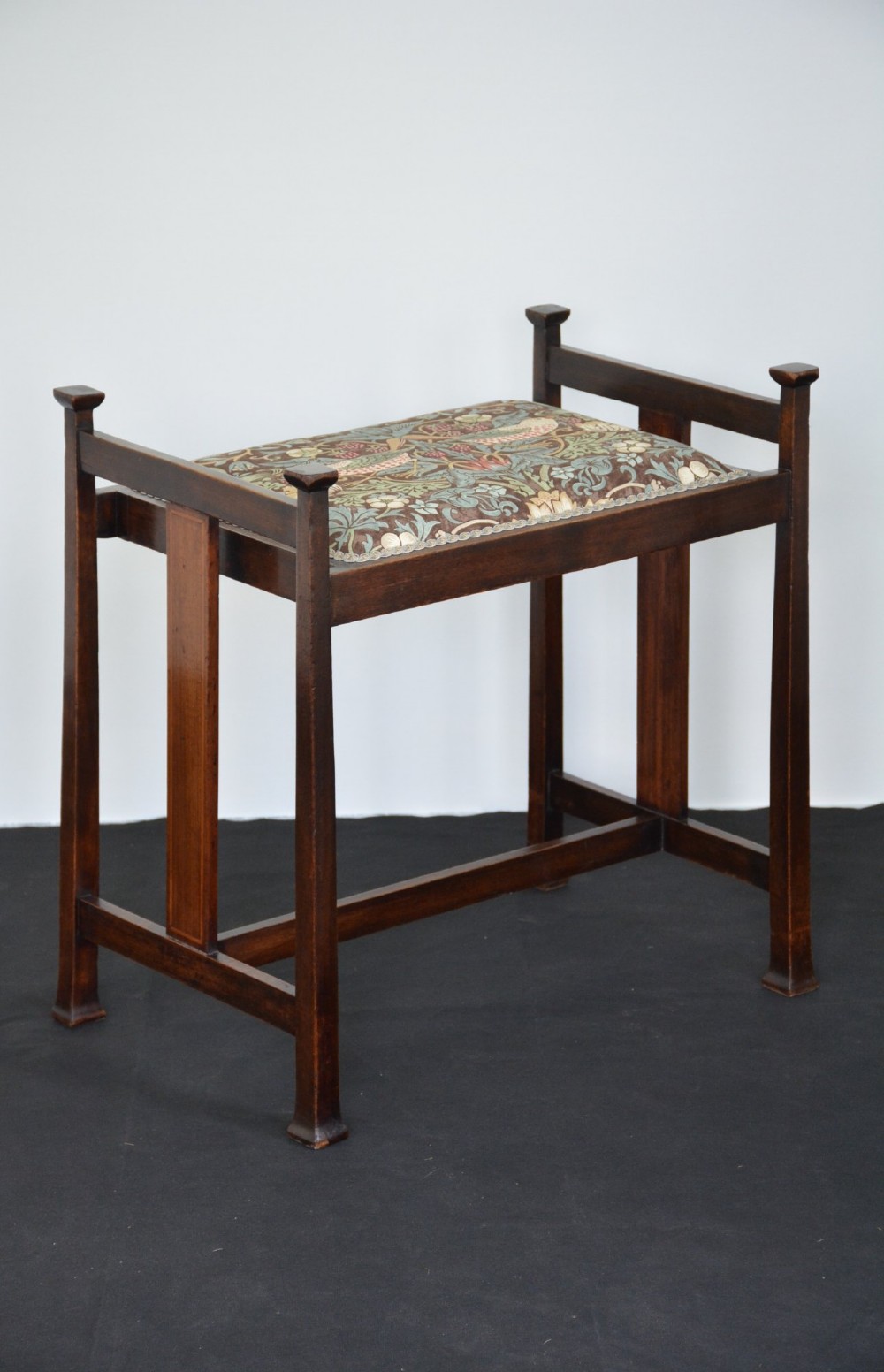 An antique dark mahogany Arts and Crafts stool featuring upright corner posts with square finials, vertical side splats, and a pattern-upholstered fabric seat.