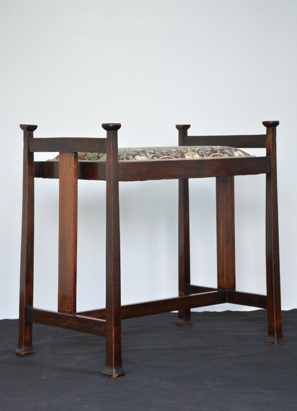 An antique dark mahogany Arts and Crafts stool featuring upright corner posts with square finials, vertical side splats, and a pattern-upholstered fabric seat. View XI