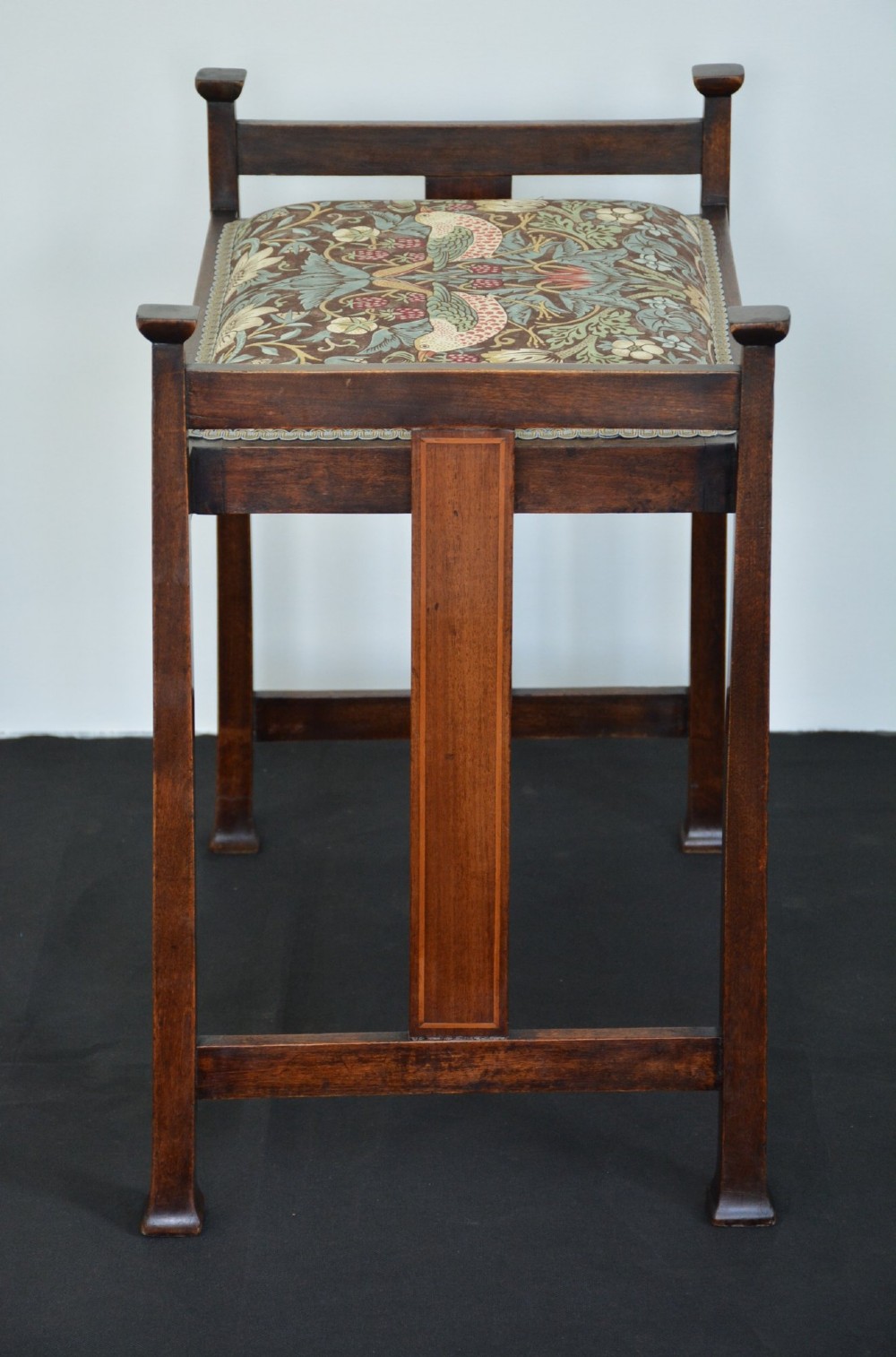 An antique dark mahogany Arts and Crafts stool featuring upright corner posts with square finials, vertical side splats, and a pattern-upholstered fabric seat. View XII