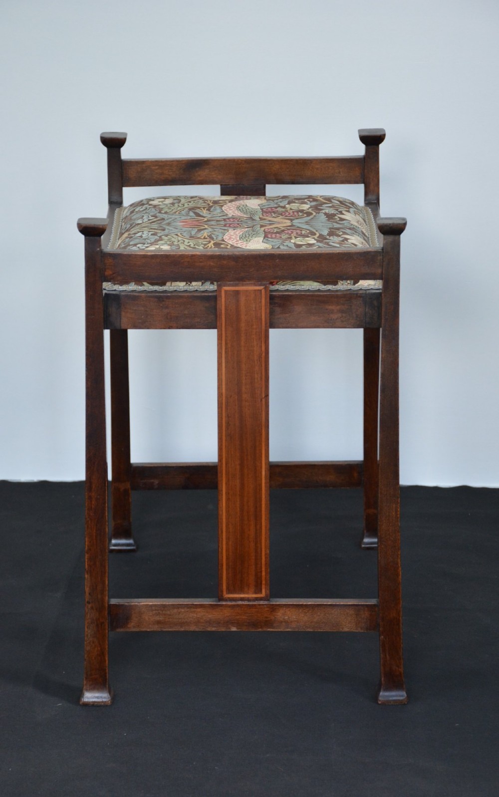 An antique dark mahogany Arts and Crafts stool featuring upright corner posts with square finials, vertical side splats, and a pattern-upholstered fabric seat. View XIV