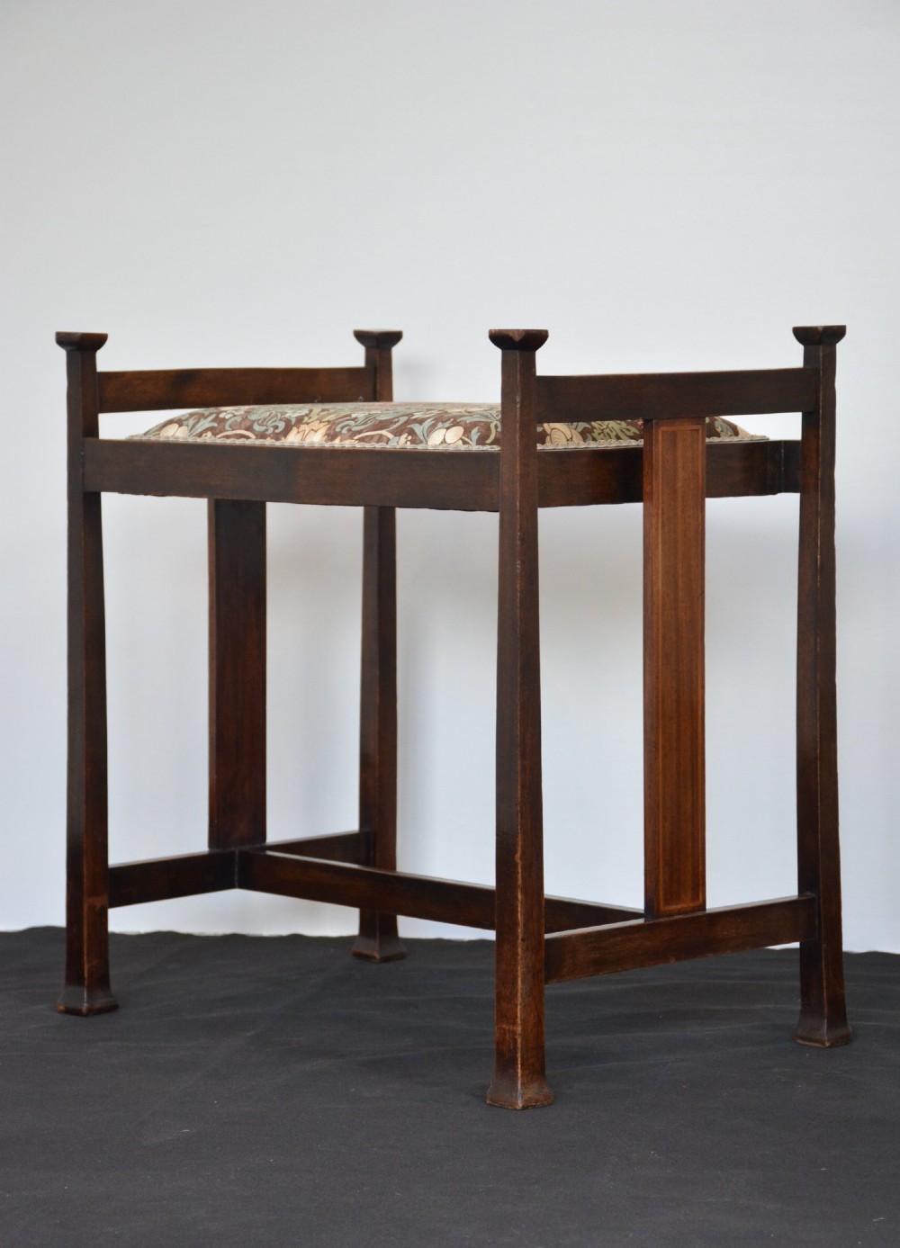 An antique dark mahogany Arts and Crafts stool featuring upright corner posts with square finials, vertical side splats, and a pattern-upholstered fabric seat. View XV