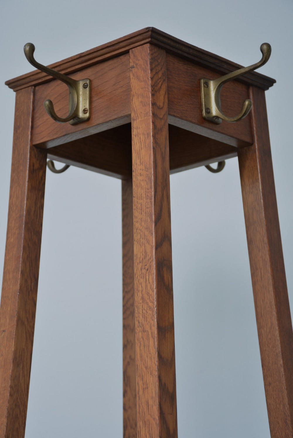 A tall solid oak freestanding coat stand featuring a tapered frame, four upper brass hooks, and a black metal drip tray at the base. View II