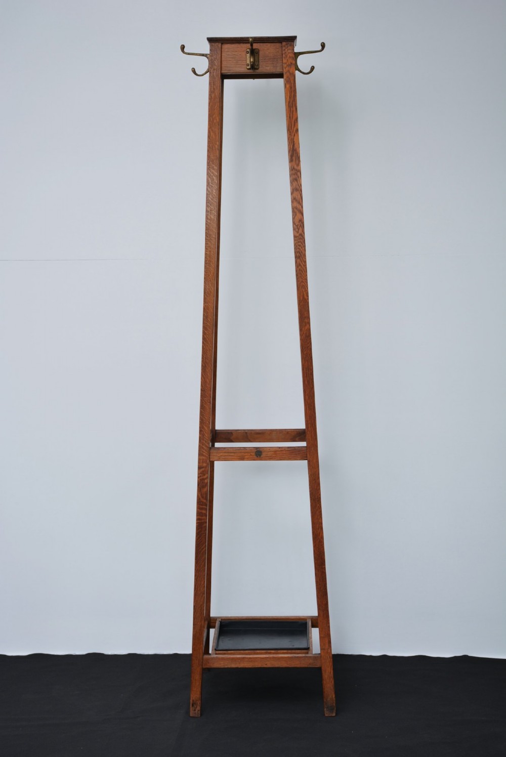 A tall solid oak freestanding coat stand featuring a tapered frame, four upper brass hooks, and a black metal drip tray at the base.