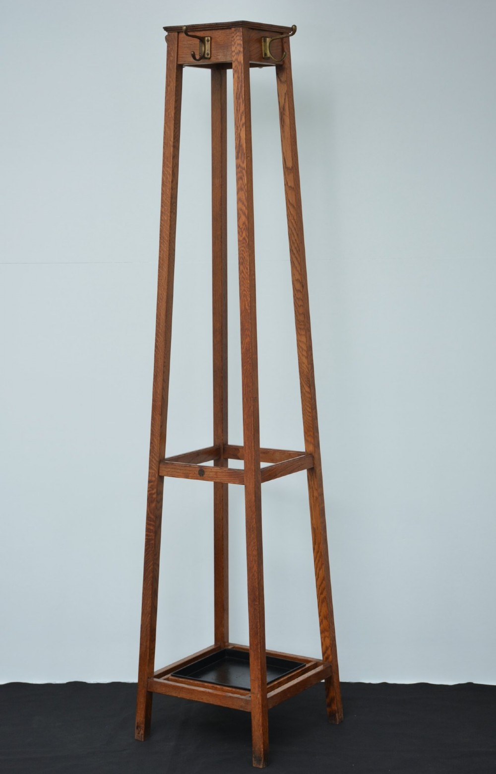 A tall solid oak freestanding coat stand featuring a tapered frame, four upper brass hooks, and a black metal drip tray at the base. View VII