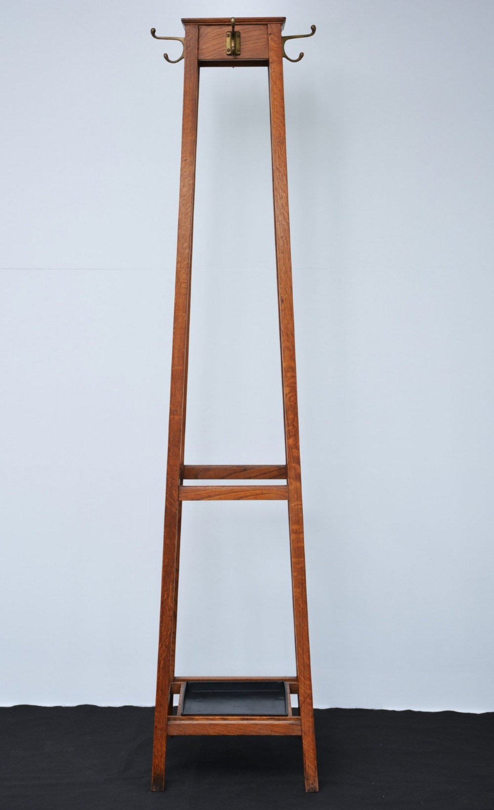 A tall solid oak freestanding coat stand featuring a tapered frame, four upper brass hooks, and a black metal drip tray at the base. View VIII