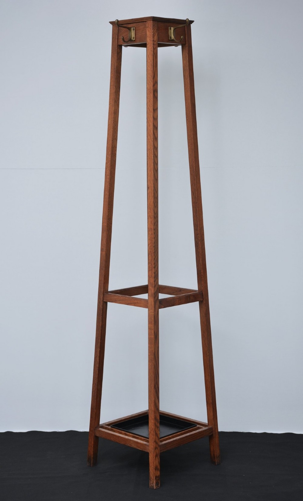 A tall solid oak freestanding coat stand featuring a tapered frame, four upper brass hooks, and a black metal drip tray at the base. View IX