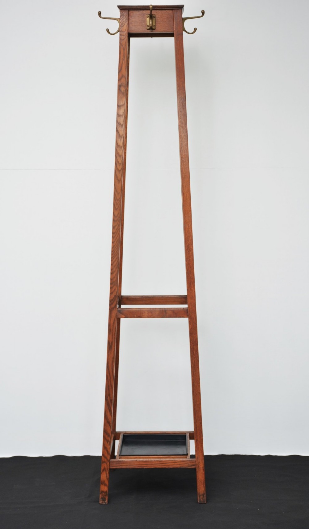A tall solid oak freestanding coat stand featuring a tapered frame, four upper brass hooks, and a black metal drip tray at the base. View X