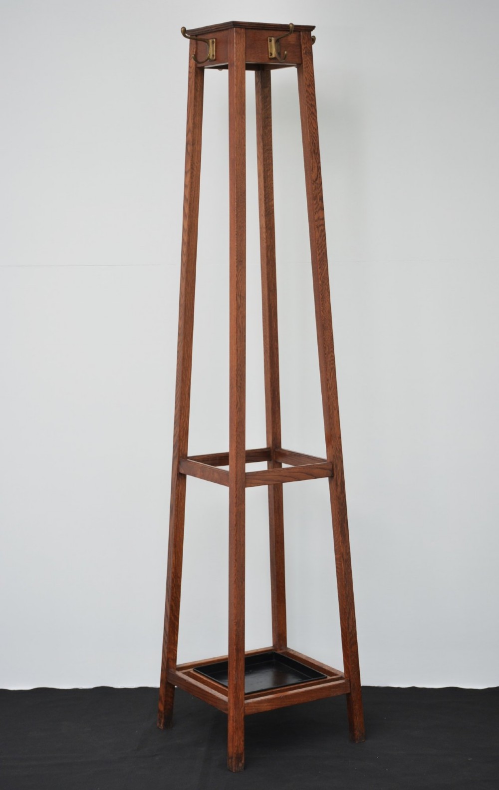 A tall solid oak freestanding coat stand featuring a tapered frame, four upper brass hooks, and a black metal drip tray at the base. View XI