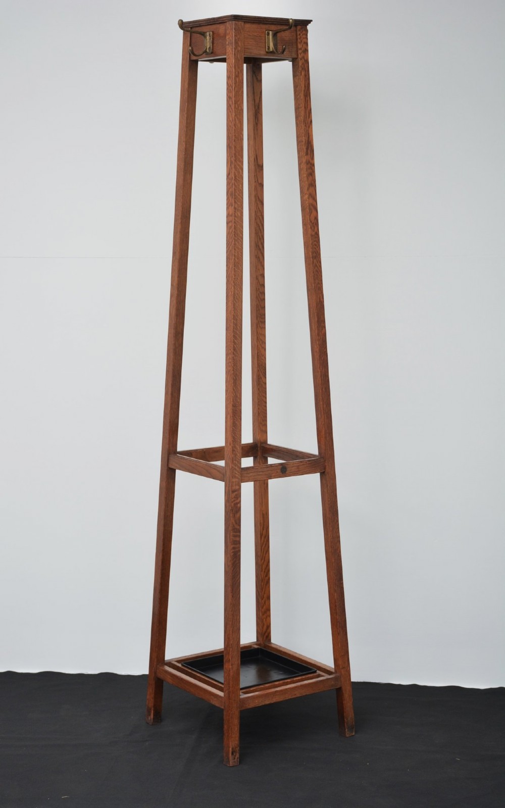 A tall solid oak freestanding coat stand featuring a tapered frame, four upper brass hooks, and a black metal drip tray at the base. View XII