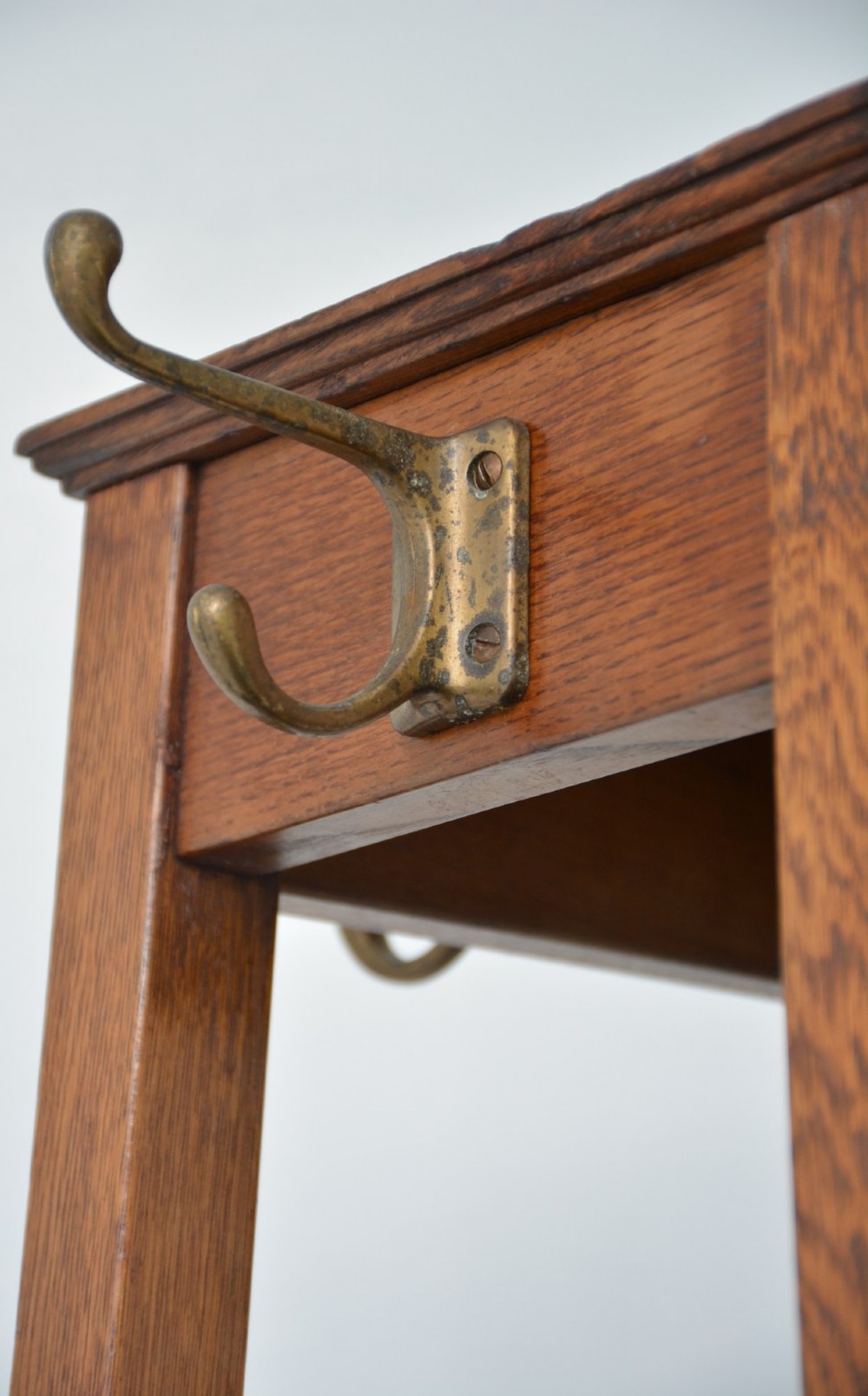 A tall solid oak freestanding coat stand featuring a tapered frame, four upper brass hooks, and a black metal drip tray at the base. View XIV