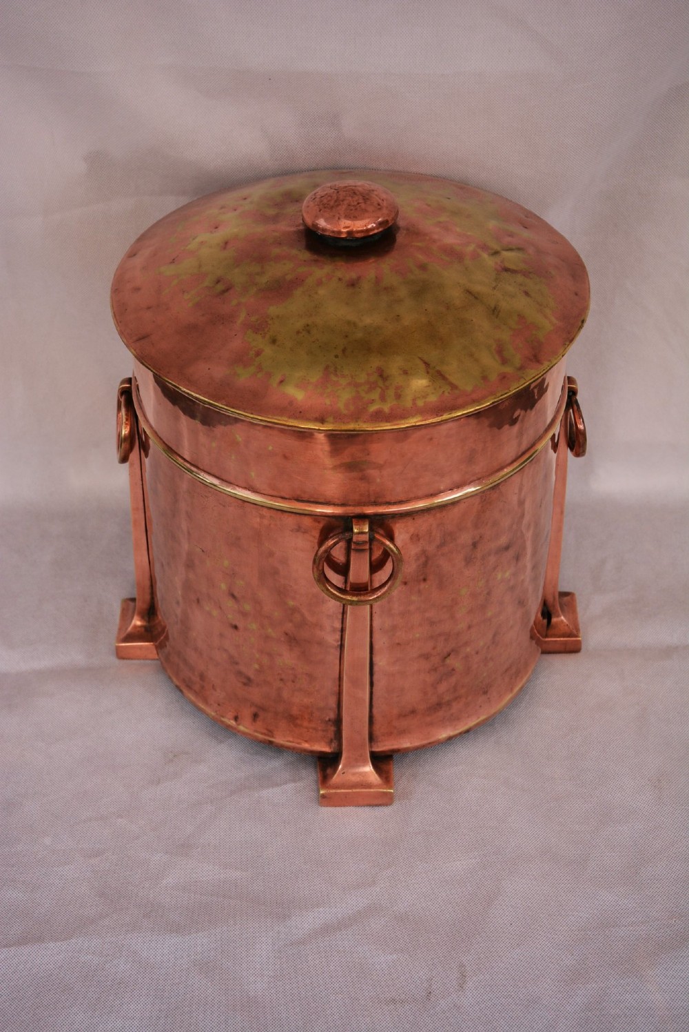 An Arts and Crafts hammered copper cylindrical coal bin featuring a fitted lid, drop side ring handles, structural legs, and a removable metal liner. View VIII