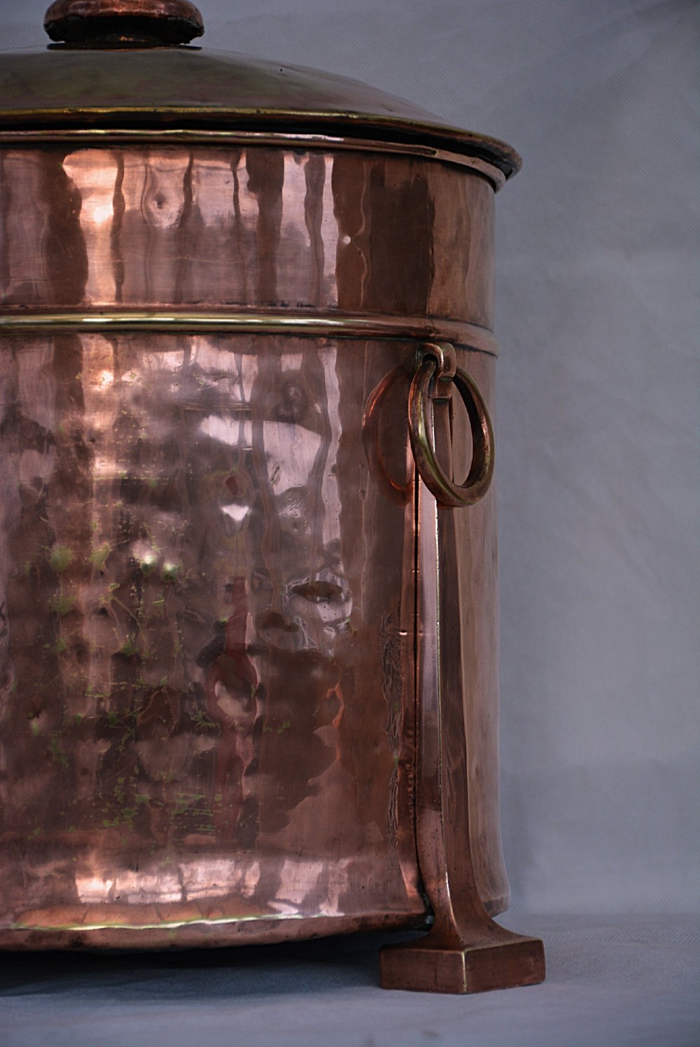 An Arts and Crafts hammered copper cylindrical coal bin featuring a fitted lid, drop side ring handles, structural legs, and a removable metal liner. View X