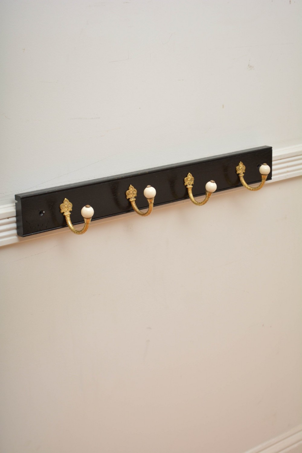 A wall-mounted black wooden coat rack featuring four decorative brass hooks with round white ceramic finials.