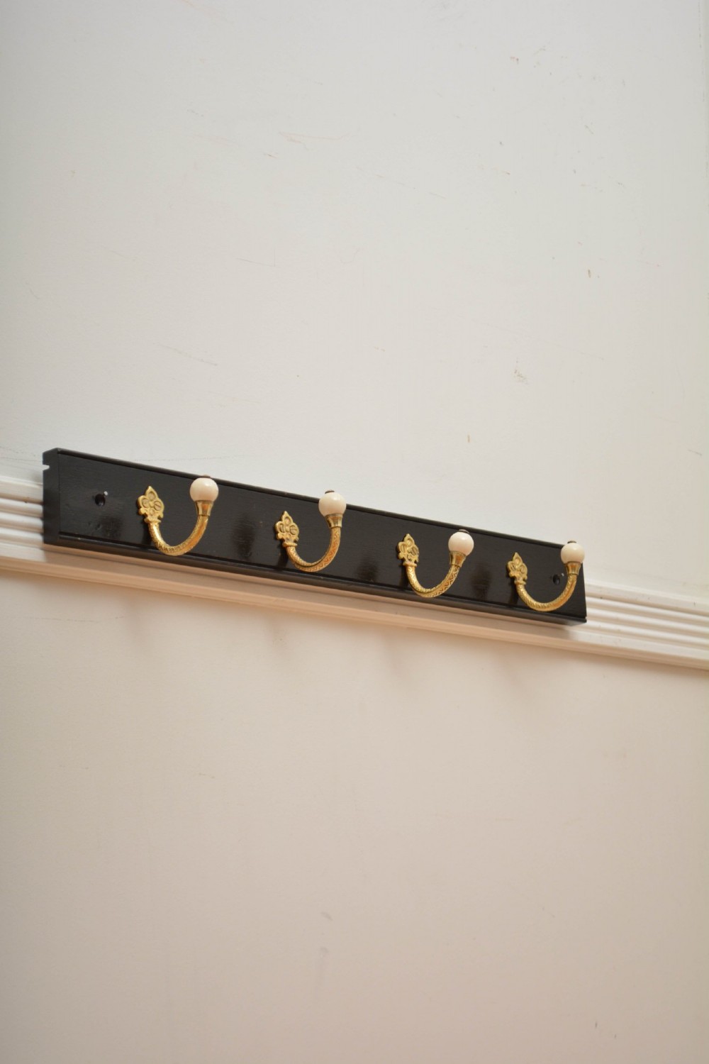 A wall-mounted black wooden coat rack featuring four decorative brass hooks with round white ceramic finials. View VI