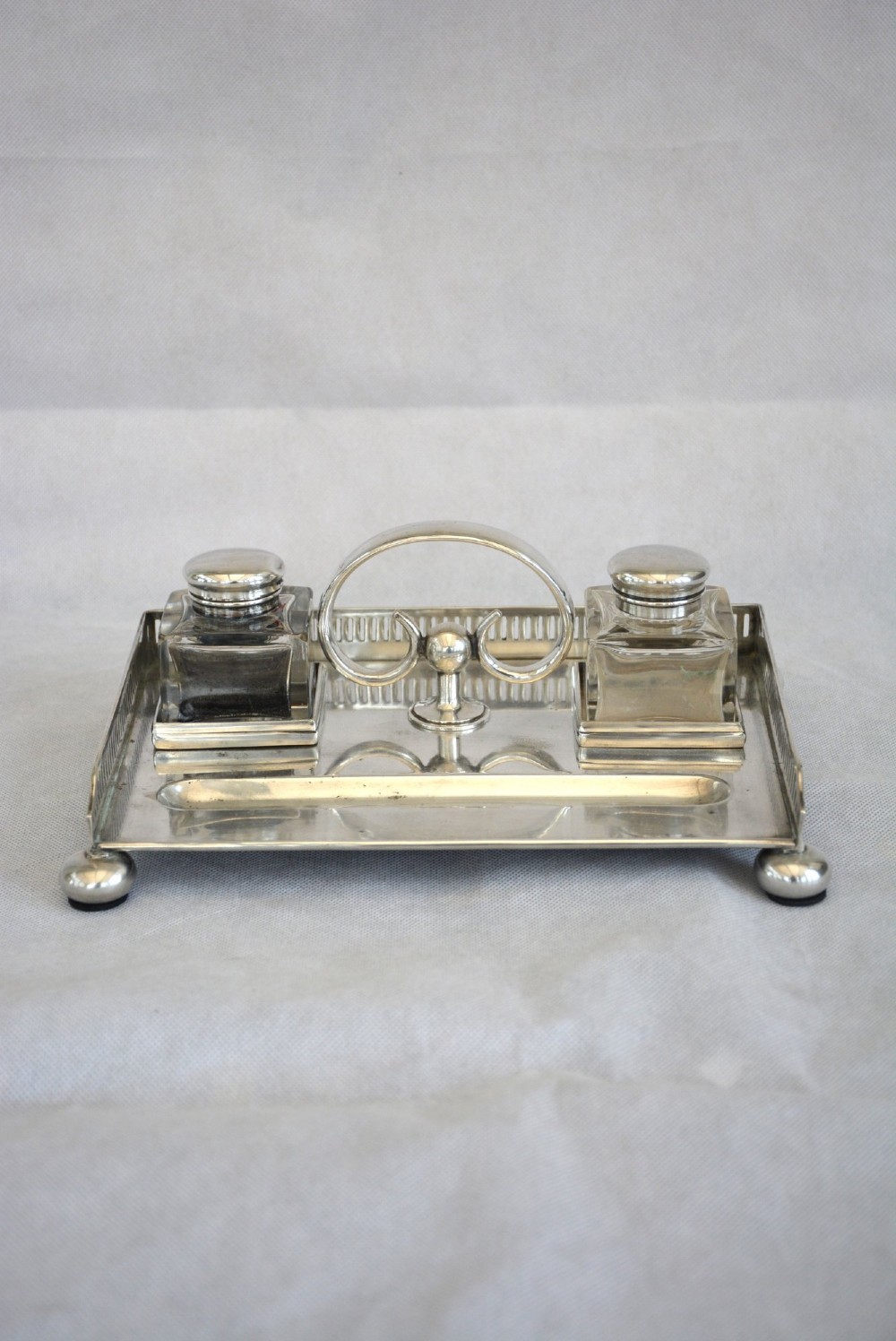 A rectangular silver-plated inkstand featuring a pierced gallery, central handle, built-in pen groove, and two clear glass inkwells with silver lids resting on ball feet. View II