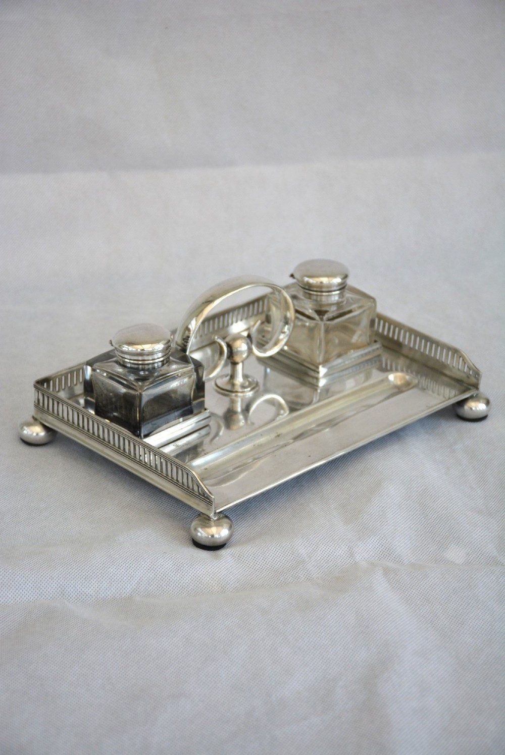 A rectangular silver-plated inkstand featuring a pierced gallery, central handle, built-in pen groove, and two clear glass inkwells with silver lids resting on ball feet. View III