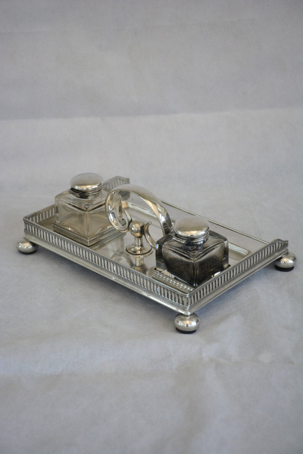 A rectangular silver-plated inkstand featuring a pierced gallery, central handle, built-in pen groove, and two clear glass inkwells with silver lids resting on ball feet. View IV