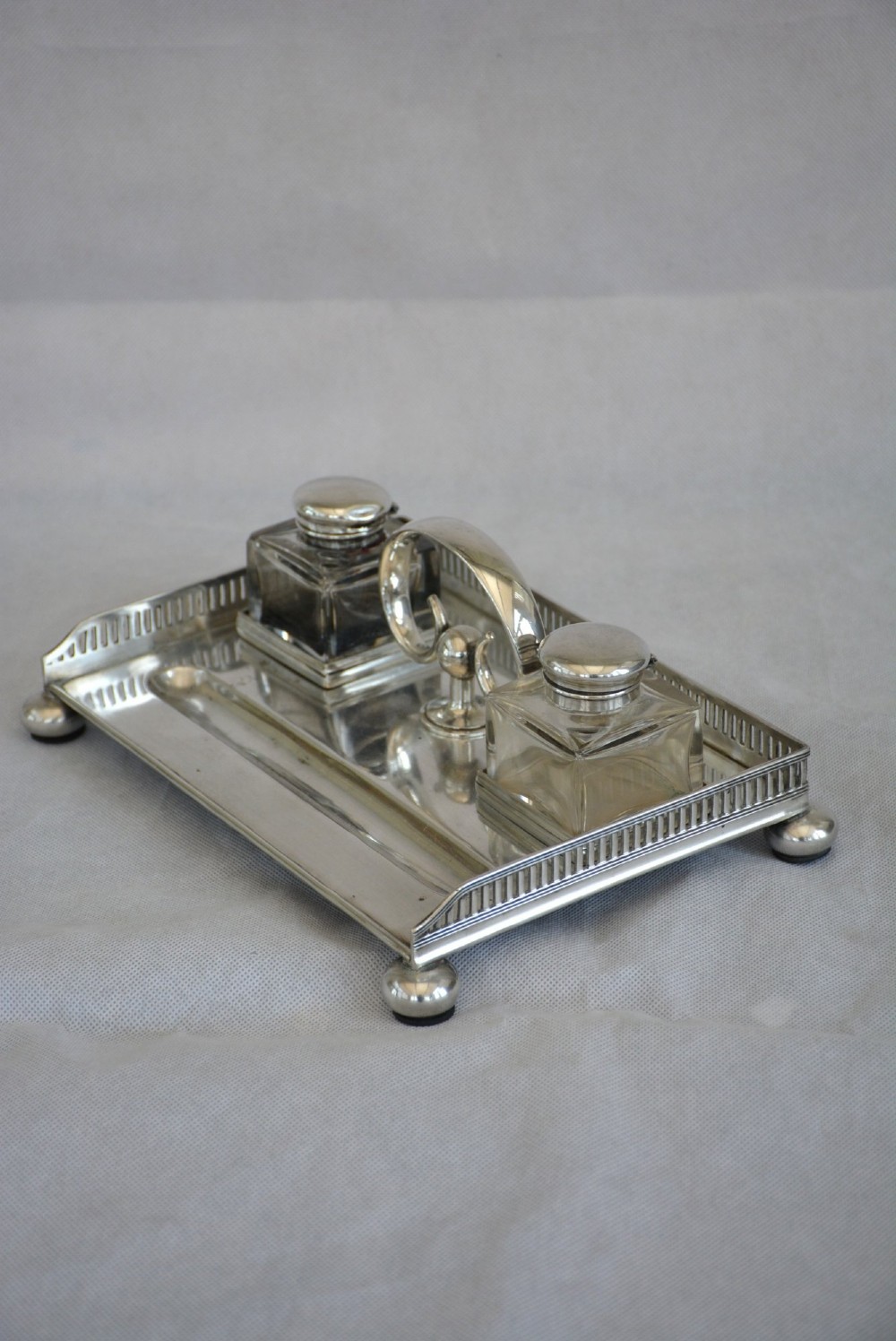 A rectangular silver-plated inkstand featuring a pierced gallery, central handle, built-in pen groove, and two clear glass inkwells with silver lids resting on ball feet. View V