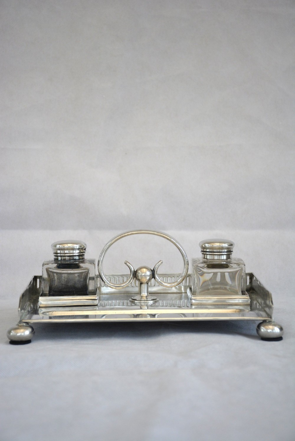 A rectangular silver-plated inkstand featuring a pierced gallery, central handle, built-in pen groove, and two clear glass inkwells with silver lids resting on ball feet. View VI