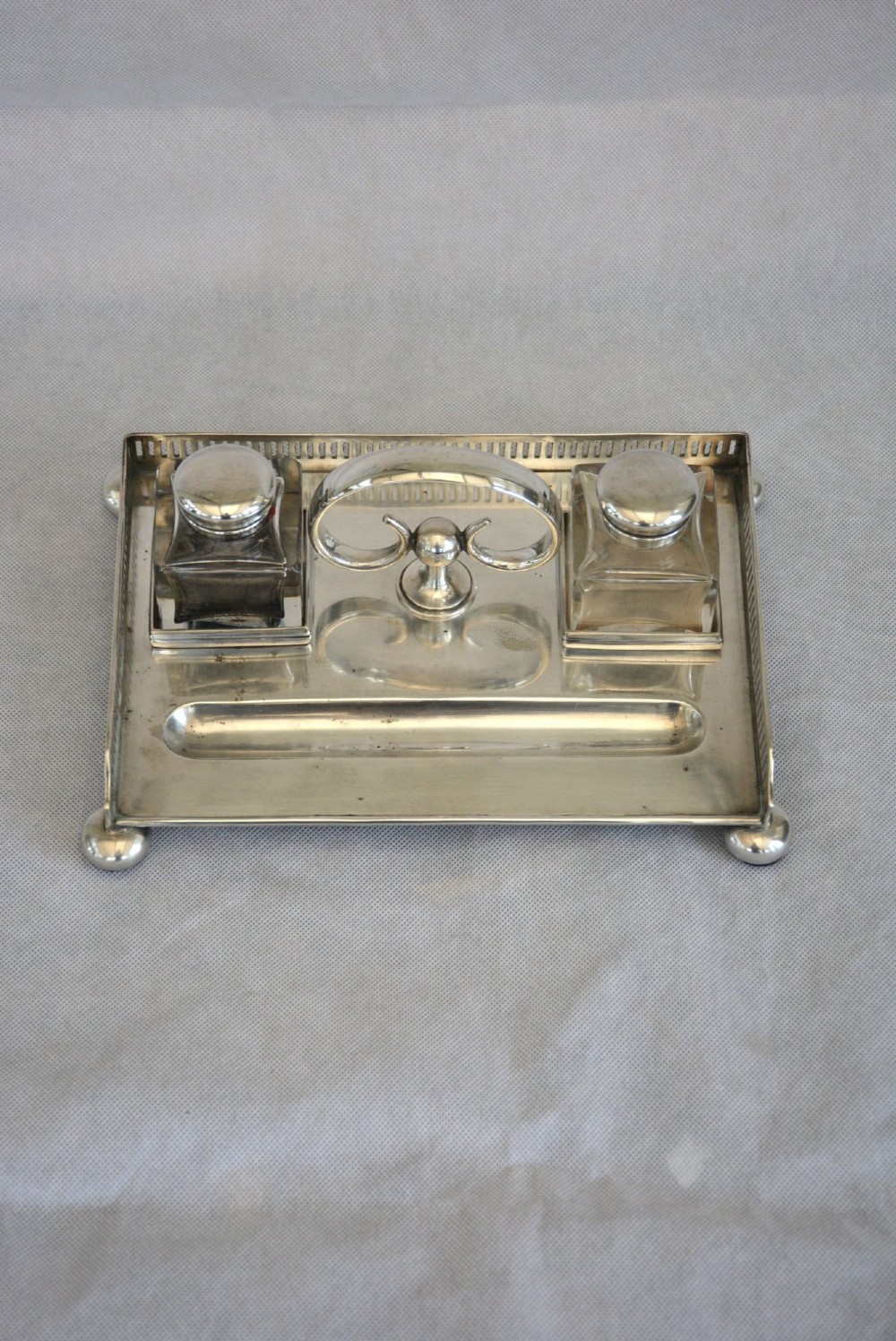 A rectangular silver-plated inkstand featuring a pierced gallery, central handle, built-in pen groove, and two clear glass inkwells with silver lids resting on ball feet. View VII