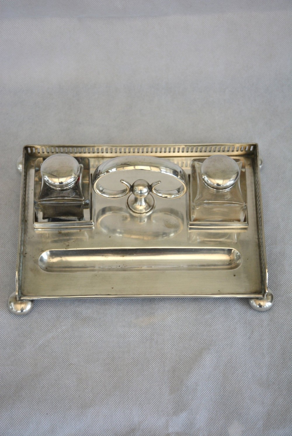 A rectangular silver-plated inkstand featuring a pierced gallery, central handle, built-in pen groove, and two clear glass inkwells with silver lids resting on ball feet. View VIII