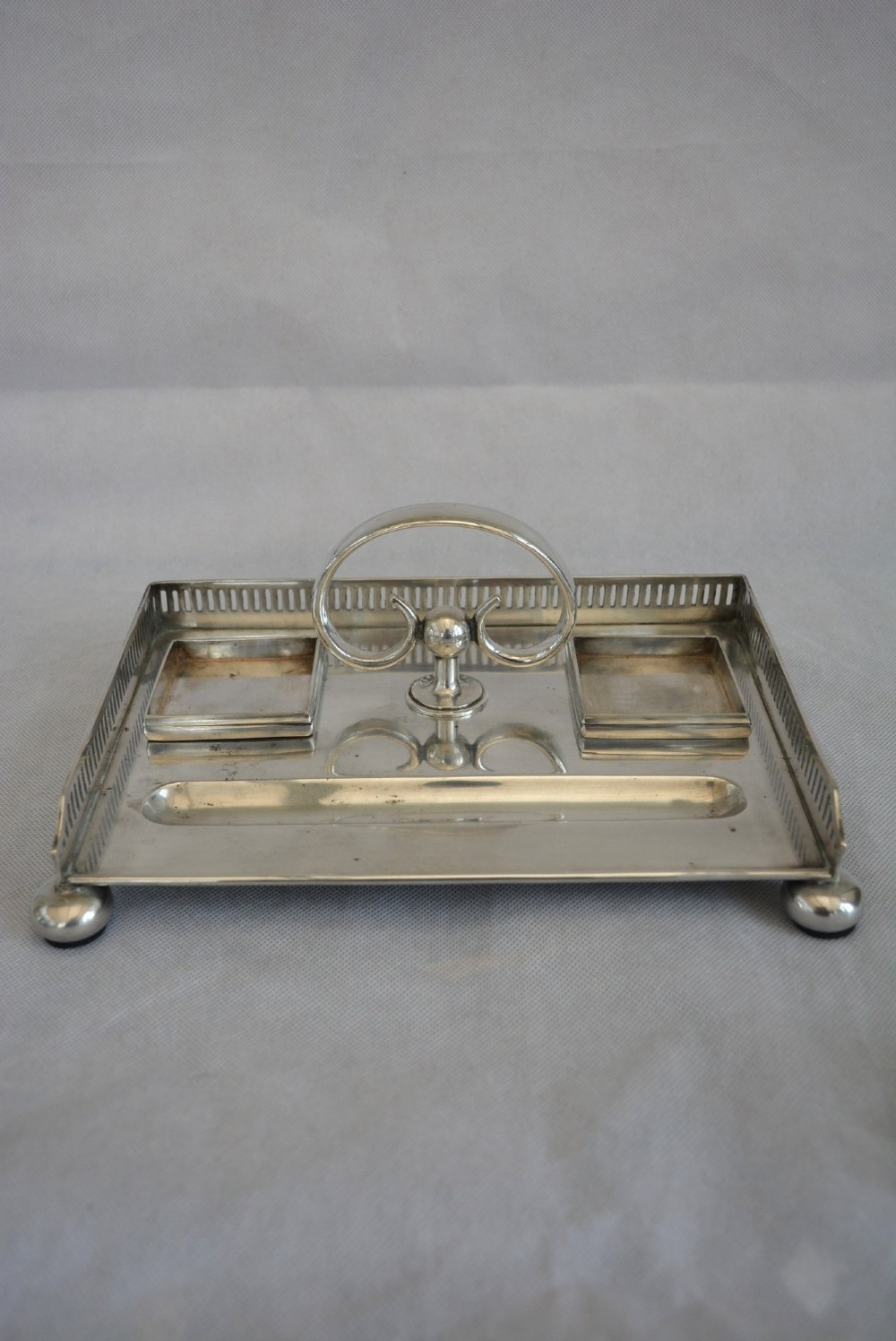 A rectangular silver-plated inkstand featuring a pierced gallery, central handle, built-in pen groove, and two clear glass inkwells with silver lids resting on ball feet. View IX