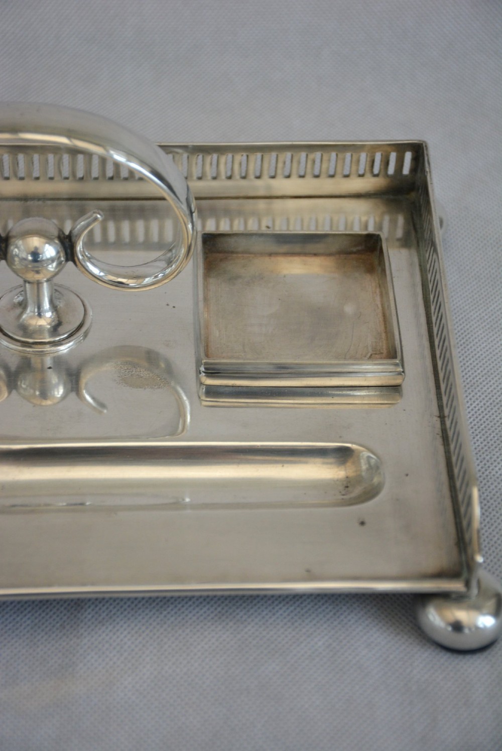 A rectangular silver-plated inkstand featuring a pierced gallery, central handle, built-in pen groove, and two clear glass inkwells with silver lids resting on ball feet. View XI