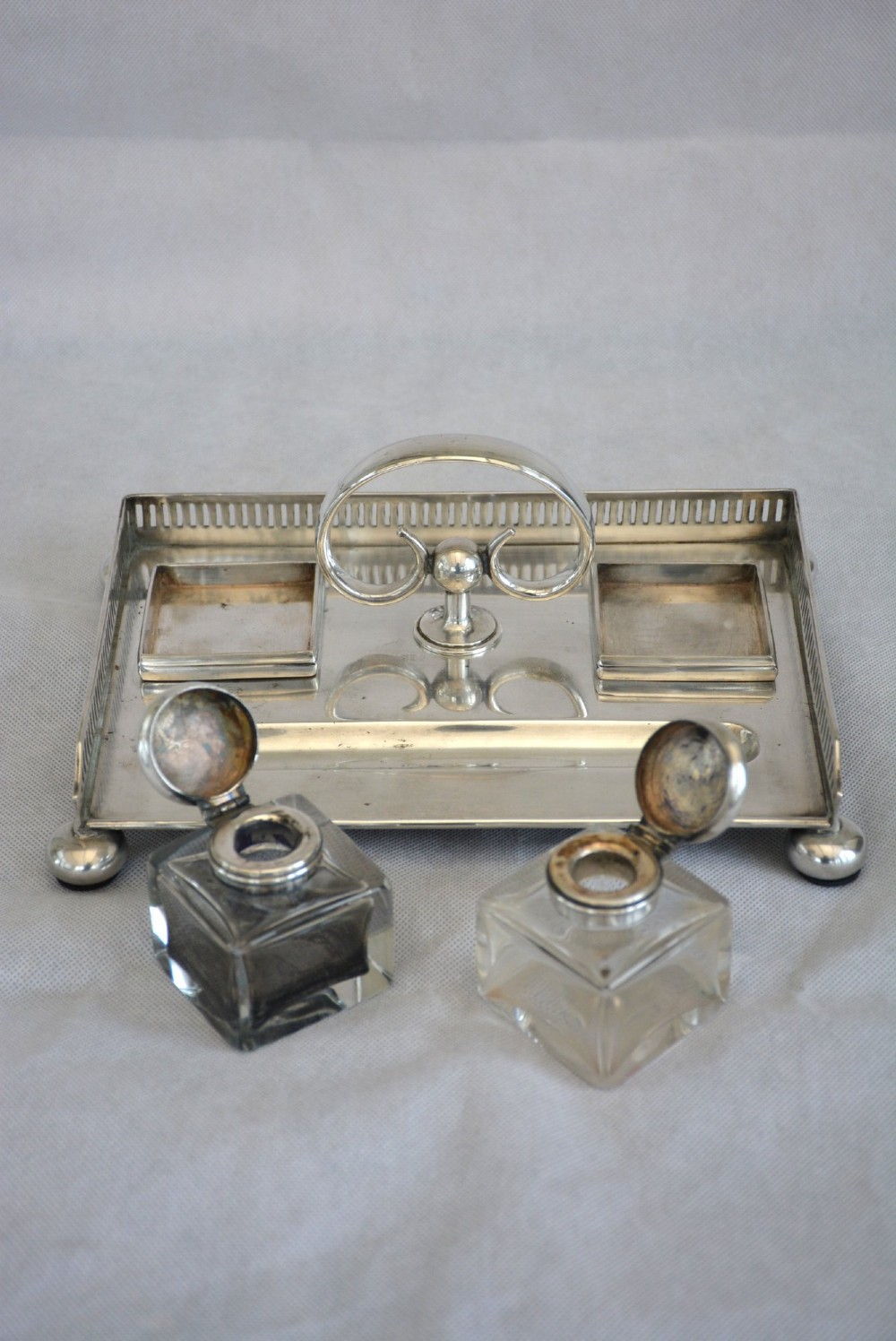 A rectangular silver-plated inkstand featuring a pierced gallery, central handle, built-in pen groove, and two clear glass inkwells with silver lids resting on ball feet. View XIII