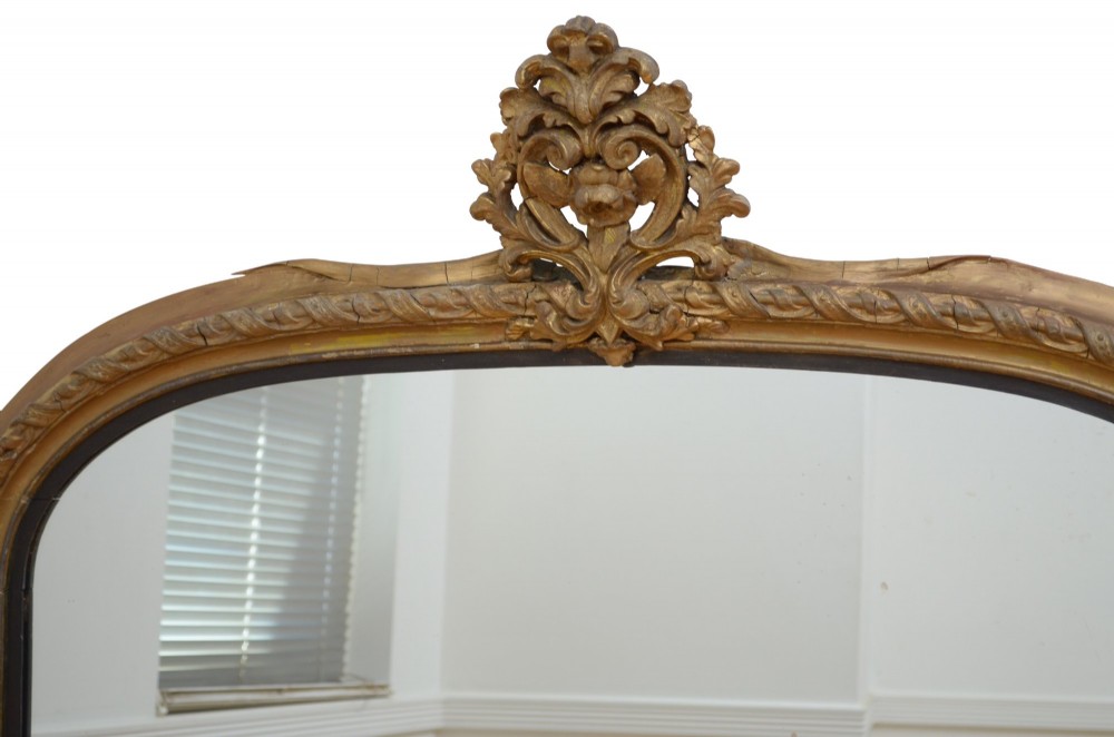 A tall arched overmantel mirror featuring a gold gilded frame decorated with an intricate carved top cresting and scroll base feet. View V