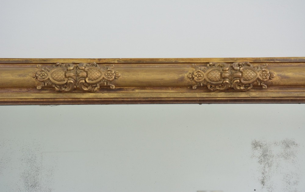 A large horizontal Regency style mirror featuring a gilded frame decorated with corner rosettes and floral mouldings enclosing foxed mirror glass. View III