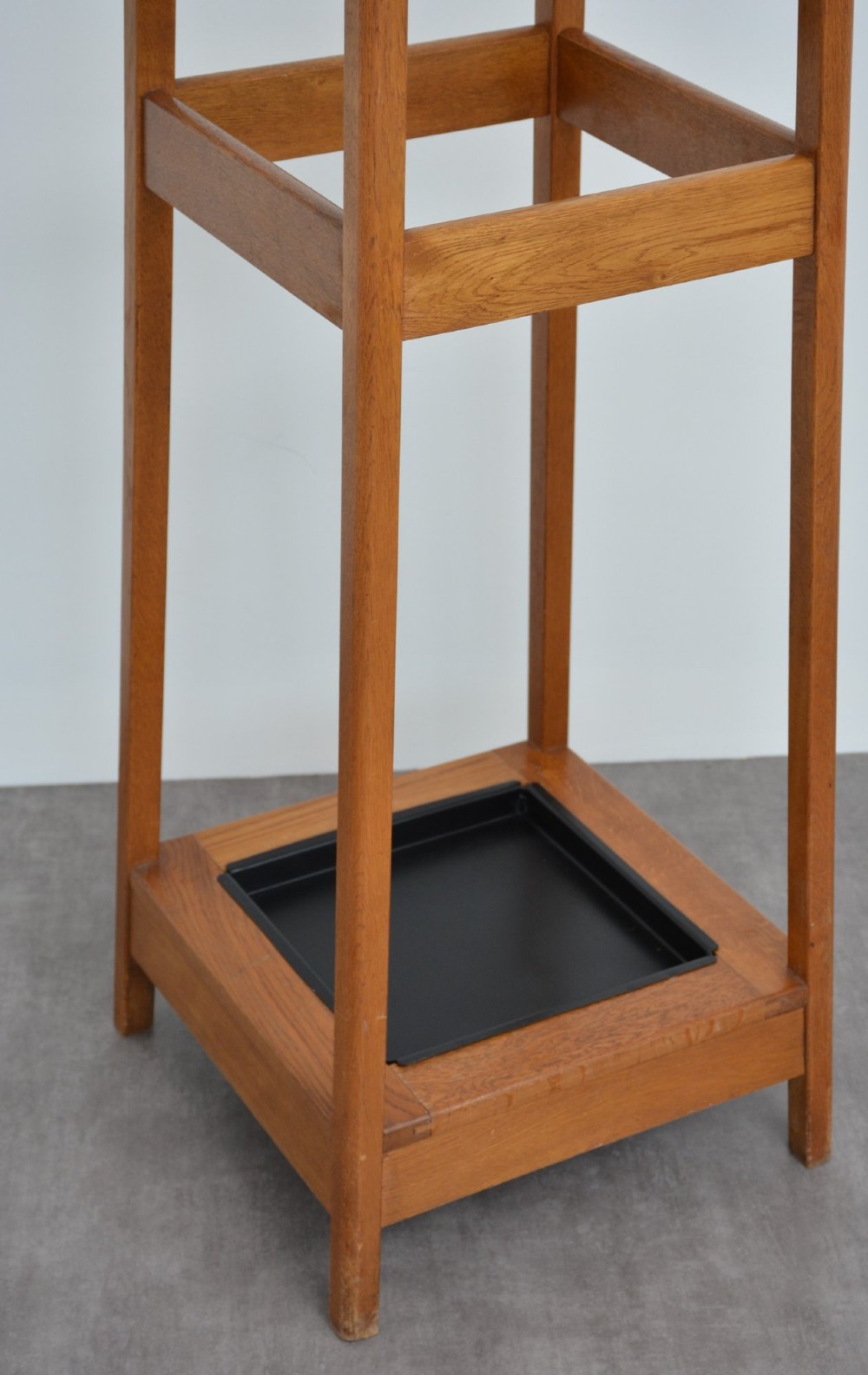 A tall, tapered oak coat stand featuring metal double hooks at the top, a square umbrella retaining rail in the centre, and a dark metal drip tray at the base. View V