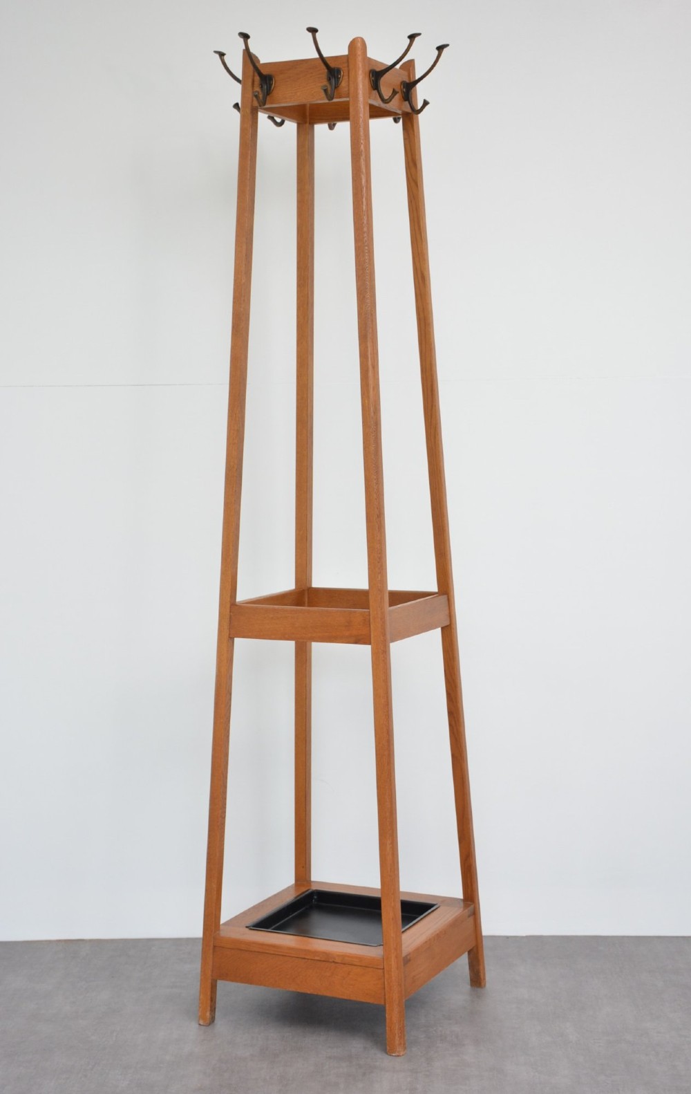 A tall, tapered oak coat stand featuring metal double hooks at the top, a square umbrella retaining rail in the centre, and a dark metal drip tray at the base.