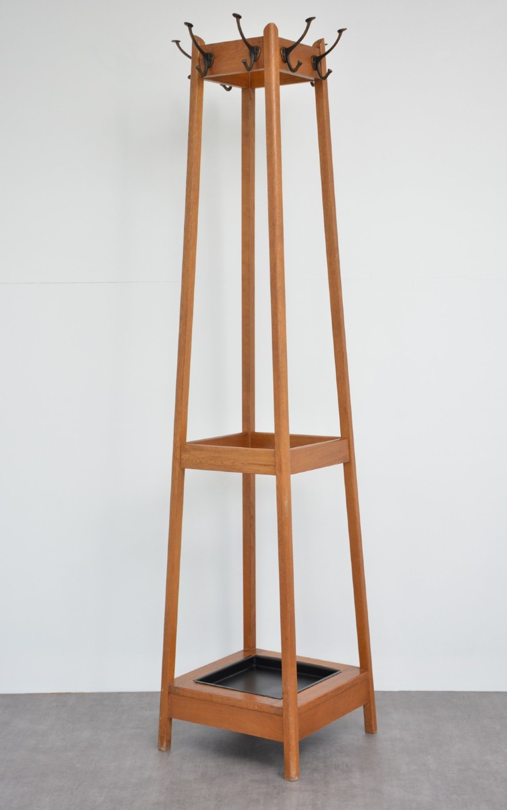 A tall, tapered oak coat stand featuring metal double hooks at the top, a square umbrella retaining rail in the centre, and a dark metal drip tray at the base. View X