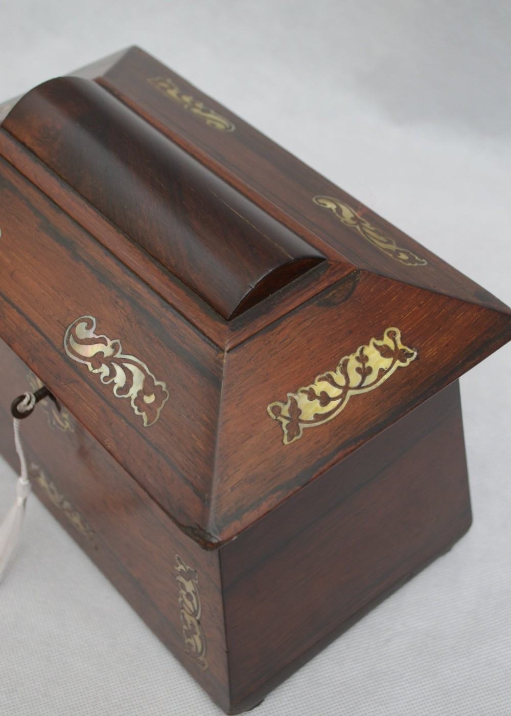 A Regency-era sarcophagus-shaped rosewood jewellery box featuring decorative scroll inlay and a key with a white tassel. View V