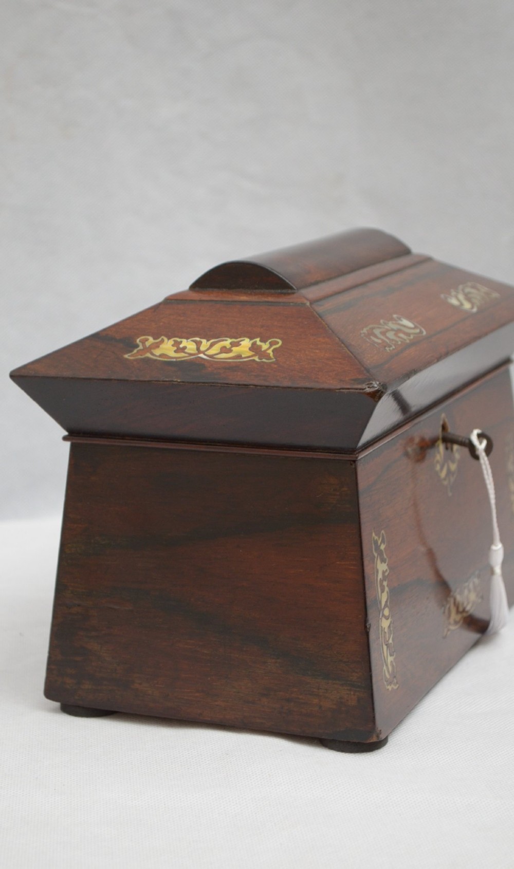 A Regency-era sarcophagus-shaped rosewood jewellery box featuring decorative scroll inlay and a key with a white tassel. View IX